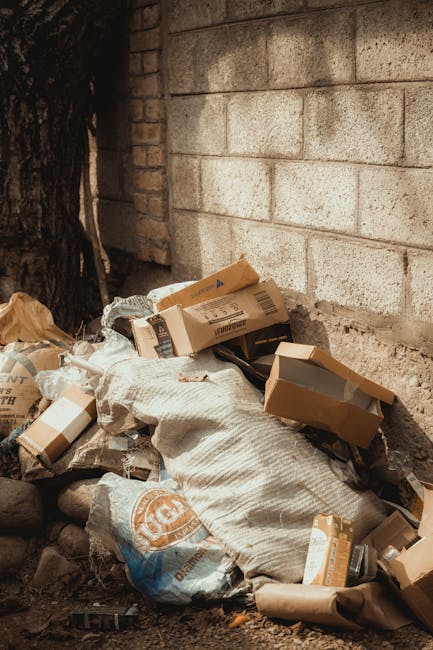 A pile of mixed waste and cardboard packaging materials situated against an outdoor brick wall with a concrete block surface. The waste includes flattened cardboard boxes, some with printed labels and