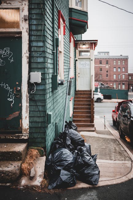 A pile of mixed waste and cardboard packaging materials situated against an outdoor brick wall with a concrete block surface. The waste includes flattened cardboard boxes, some with printed labels and