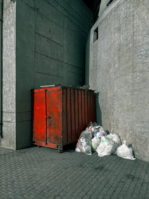 An outdoor scene showing multiple overflowing waste and rubbish bins situated on a paved sidewalk beside a curb, with various types of discarded household and mixed paper waste piled around the contai